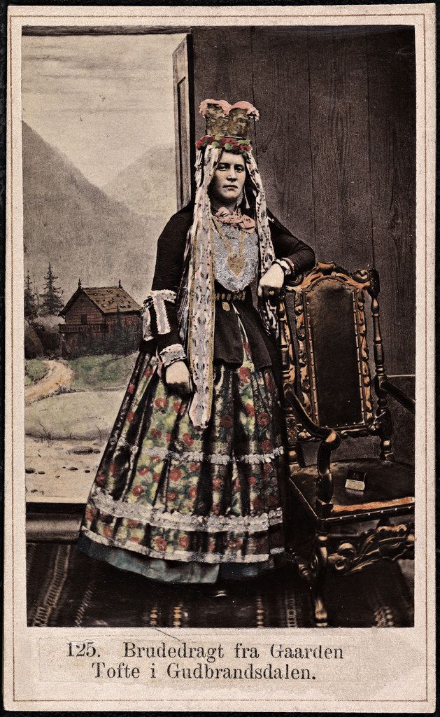 Vintage photograph of a woman in traditional Norwegian bridal attire, standing in an interior space with a carved wooden chair and a view of a landscape with a traditional wooden building and mountains.