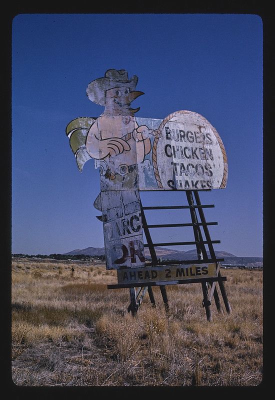 A weathered roadside sign featuring a cartoon chicken wearing a cowboy hat, advertising burgers, chicken, tacos, and shakes. The sign is located in a field with rolling hills and mountains in the background.
