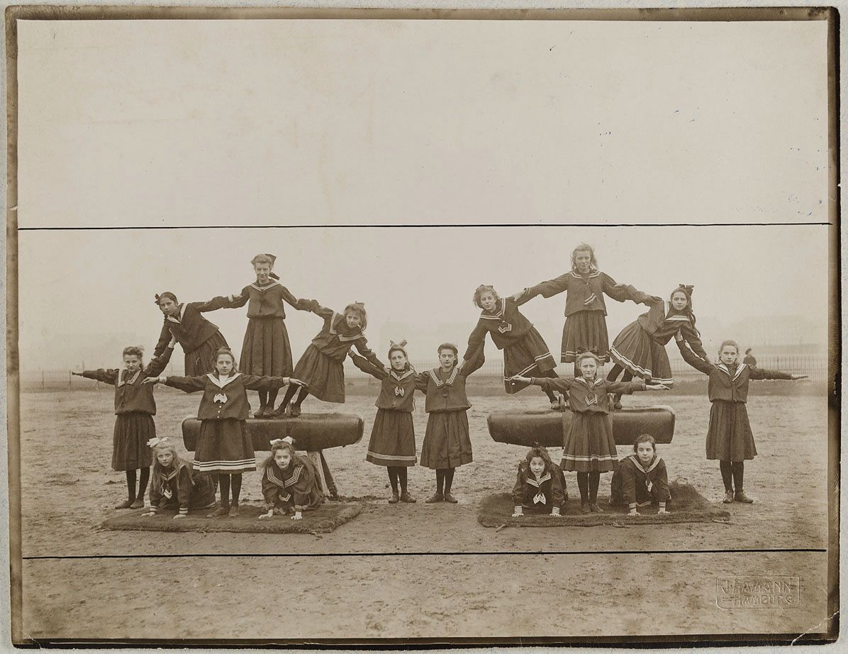 A vintage photograph of a group of young girls in uniforms posing around wooden barrels in an outdoor field.