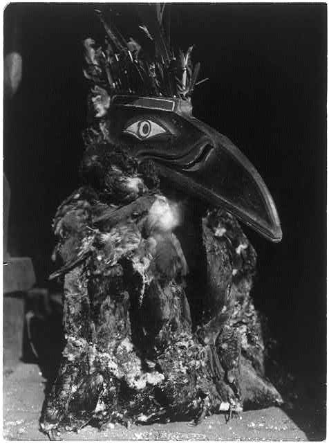 A black and white photograph of a person wearing a bird-like mask and feathered costume.