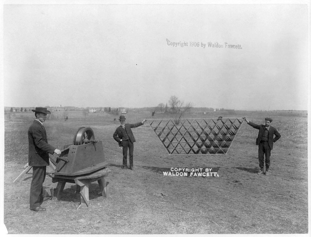 A vintage black and white photograph from 1906 showing three men in a field with a large, unusual kite structure.