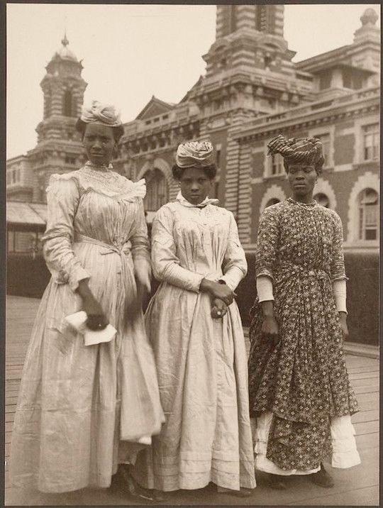 Vintage photograph of three African American women in long dresses and hats standing in front of a grand building.