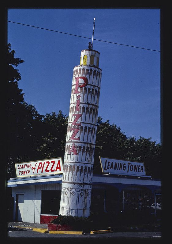 A vintage roadside attraction, the "Leaning Tower of Pizza", featuring a tall, cylindrical structure resembling the Leaning Tower of Pisa, with a low, white building and signs below.