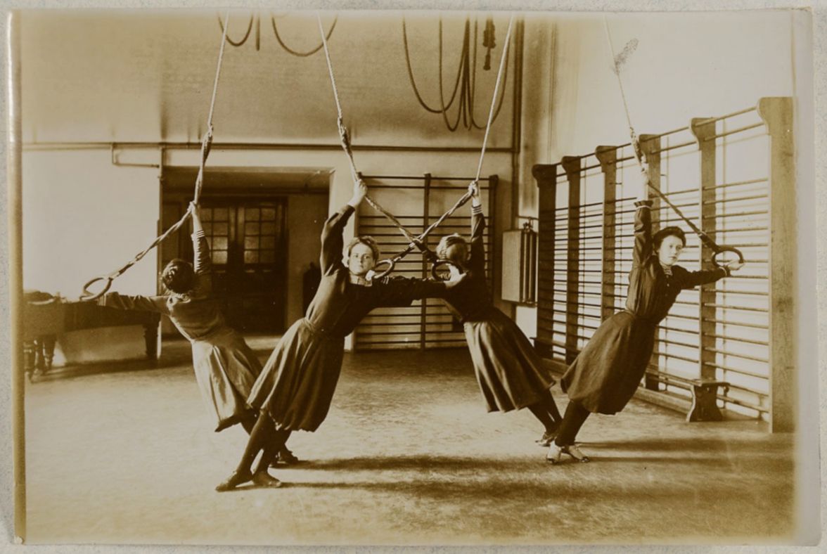 A sepia-toned photograph of four nuns in a gymnasium, suspended in the air by ropes as part of a physical exercise.