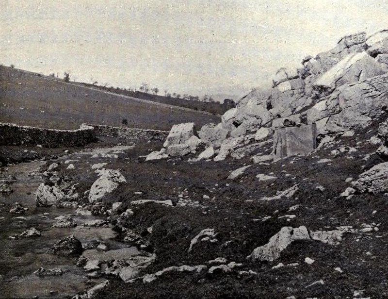 A black and white photograph of a rocky landscape with a stream, a tent, and a stone wall.