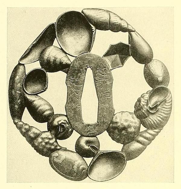 A detailed, antique circular ornament made of metal with bulbous and shell-like elements surrounding a central oval plate with engraved characters.