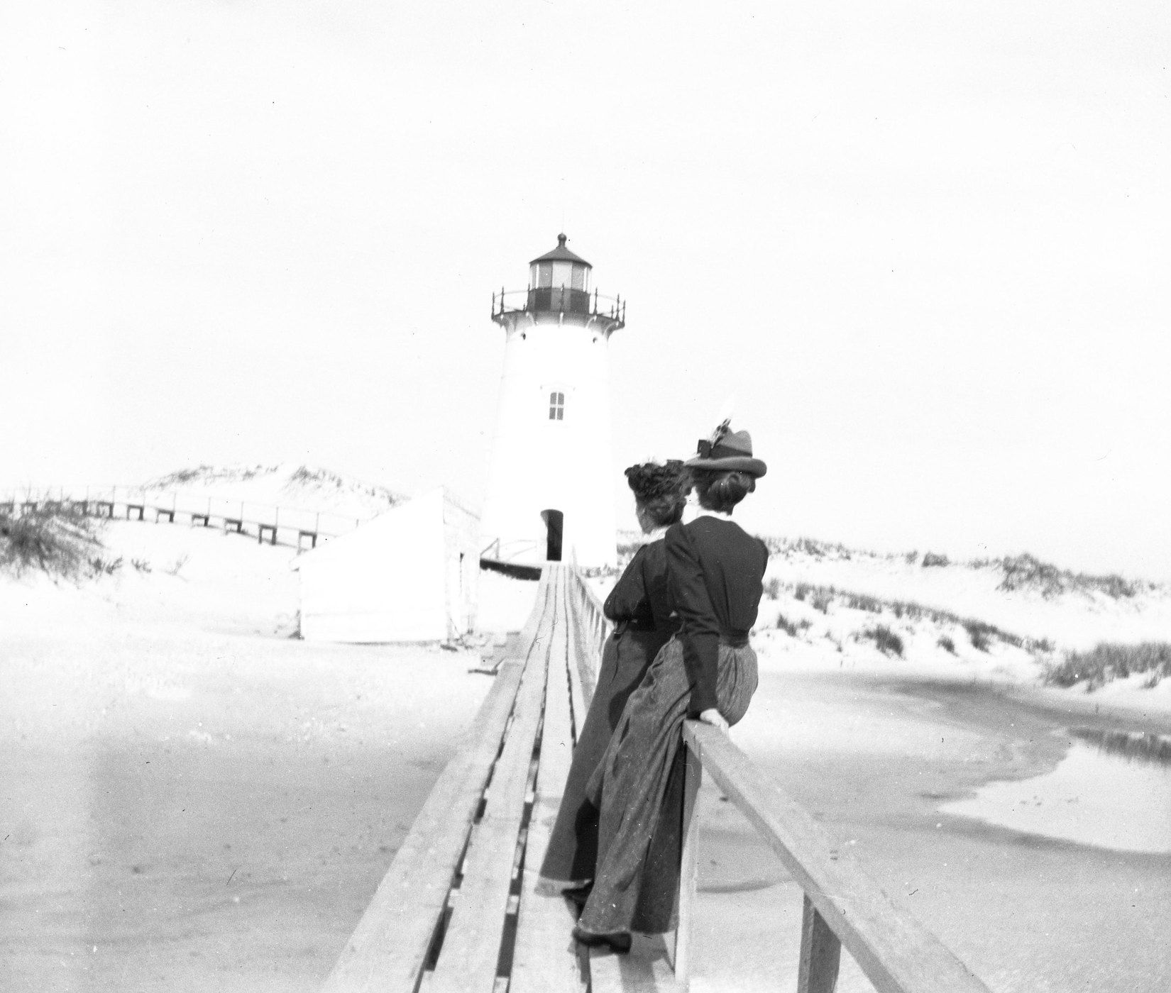 Two women stand on a wooden walkway looking towards a lighthouse in a snowy landscape.