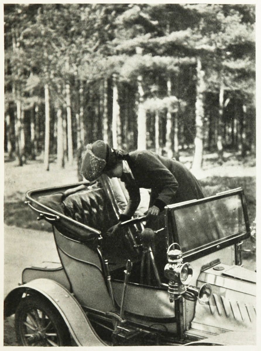 A vintage black and white photograph of a person driving an early 20th-century automobile in a wooded area.