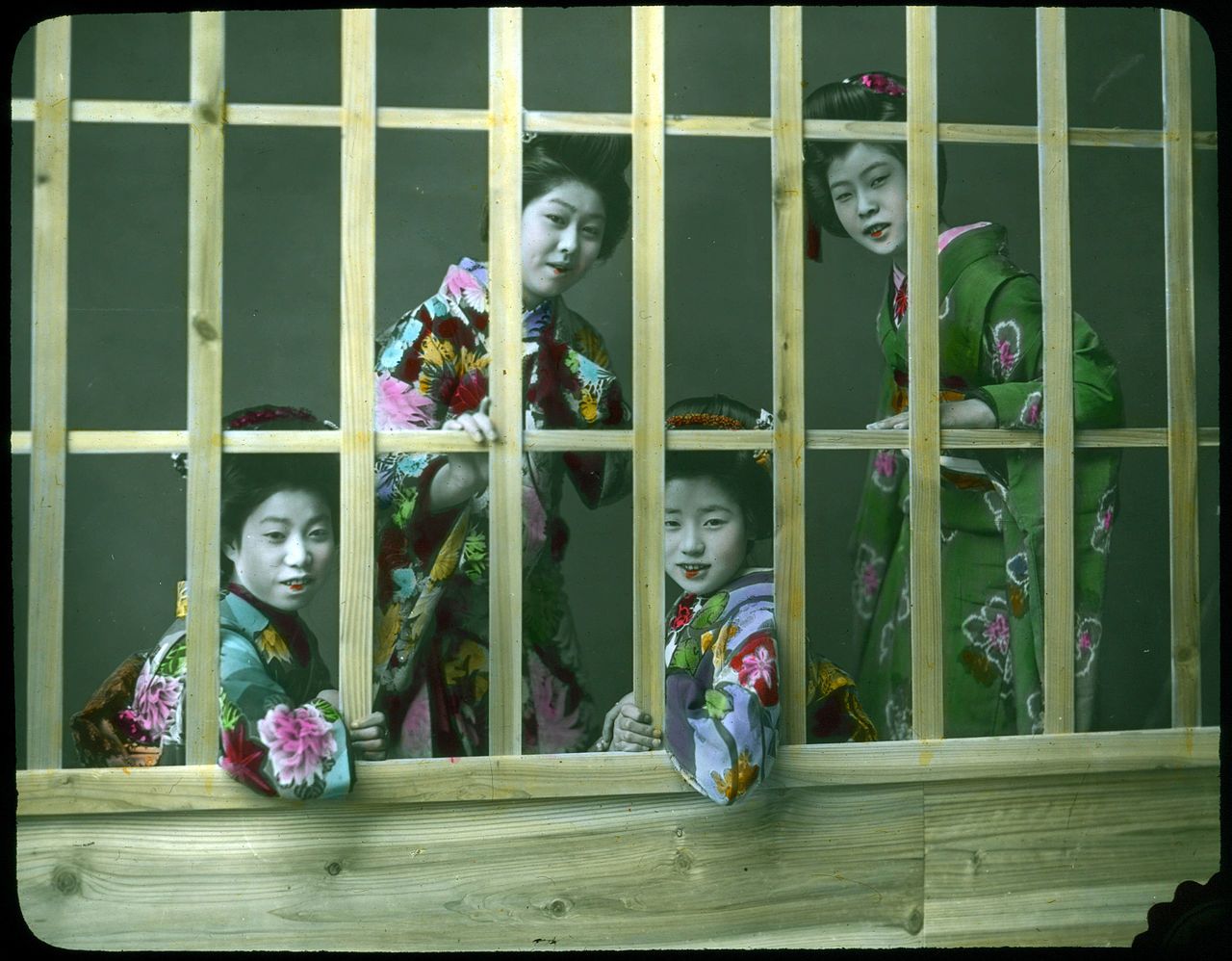 Four young Japanese girls in kimonos peer through the slats of a wooden window.
