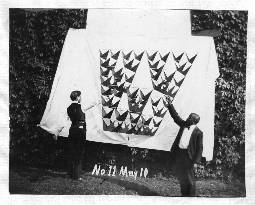 A black and white photograph of two people displaying a large quilt outdoors. The quilt features a geometric pattern of dark and light triangles. The background consists of lush greenery, with trees and grass surrounding the scene. At the bottom of the image, there is text that reads 'No 11 May 10'.