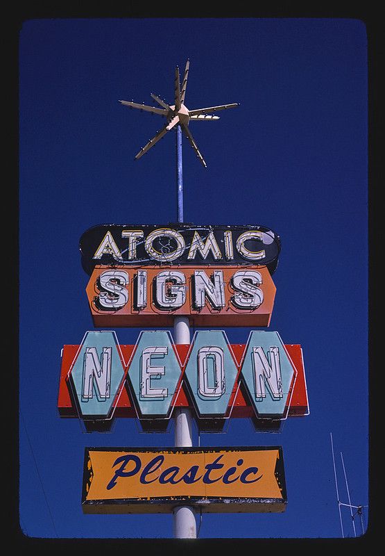 A vintage neon sign with the words 'ATOMIC', 'SIGNS', 'NEON', and 'Plastic' against a deep blue sky.
