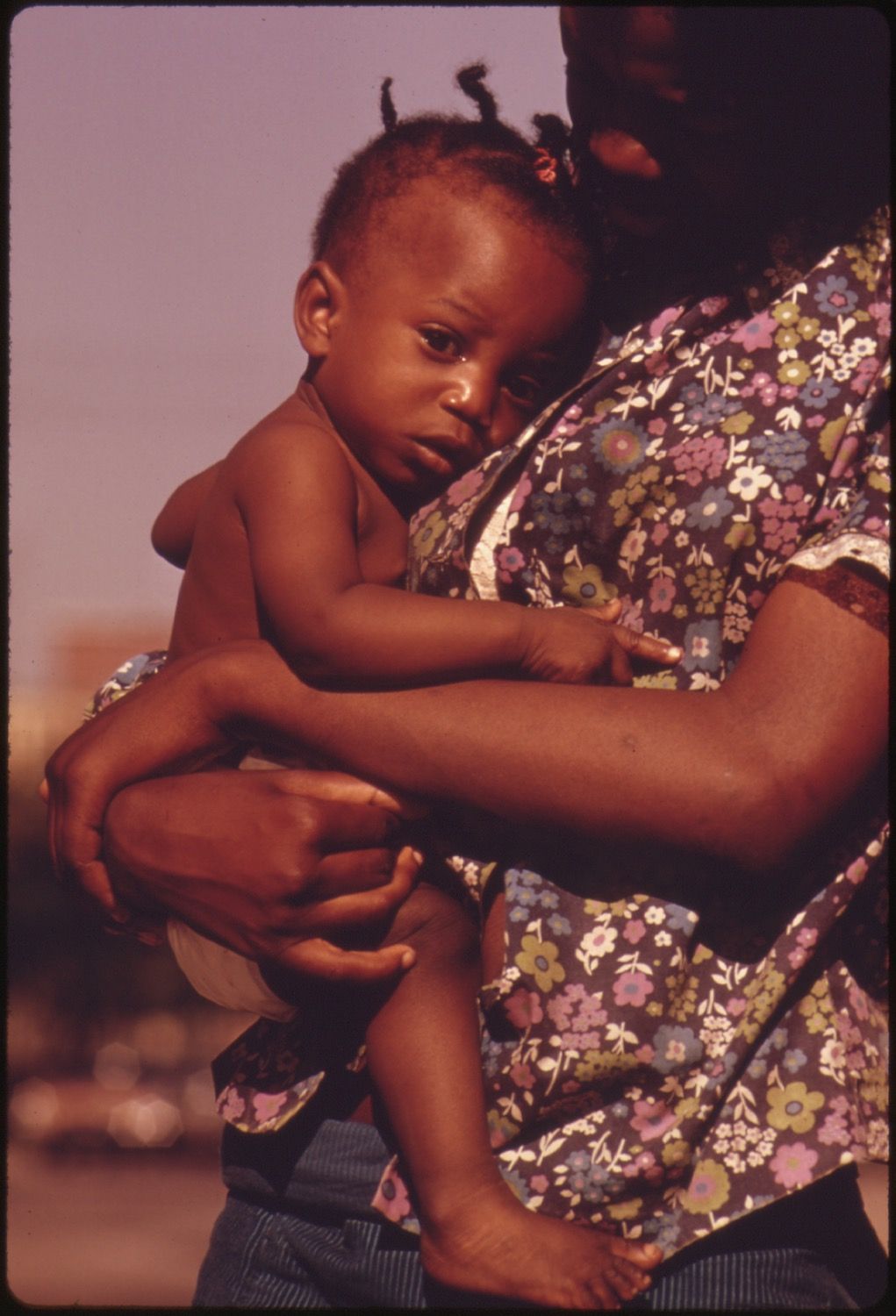 A close-up of a baby being held by an adult, with a somber mood and a blurred outdoor background.