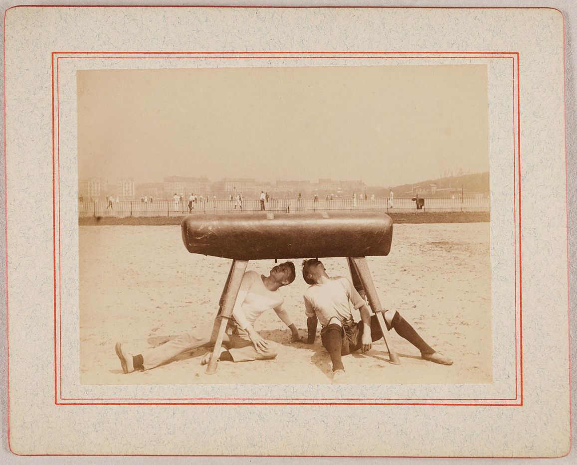 A sepia-toned vintage photograph of a gymnast performing on a vaulting horse in front of a crowd of spectators.