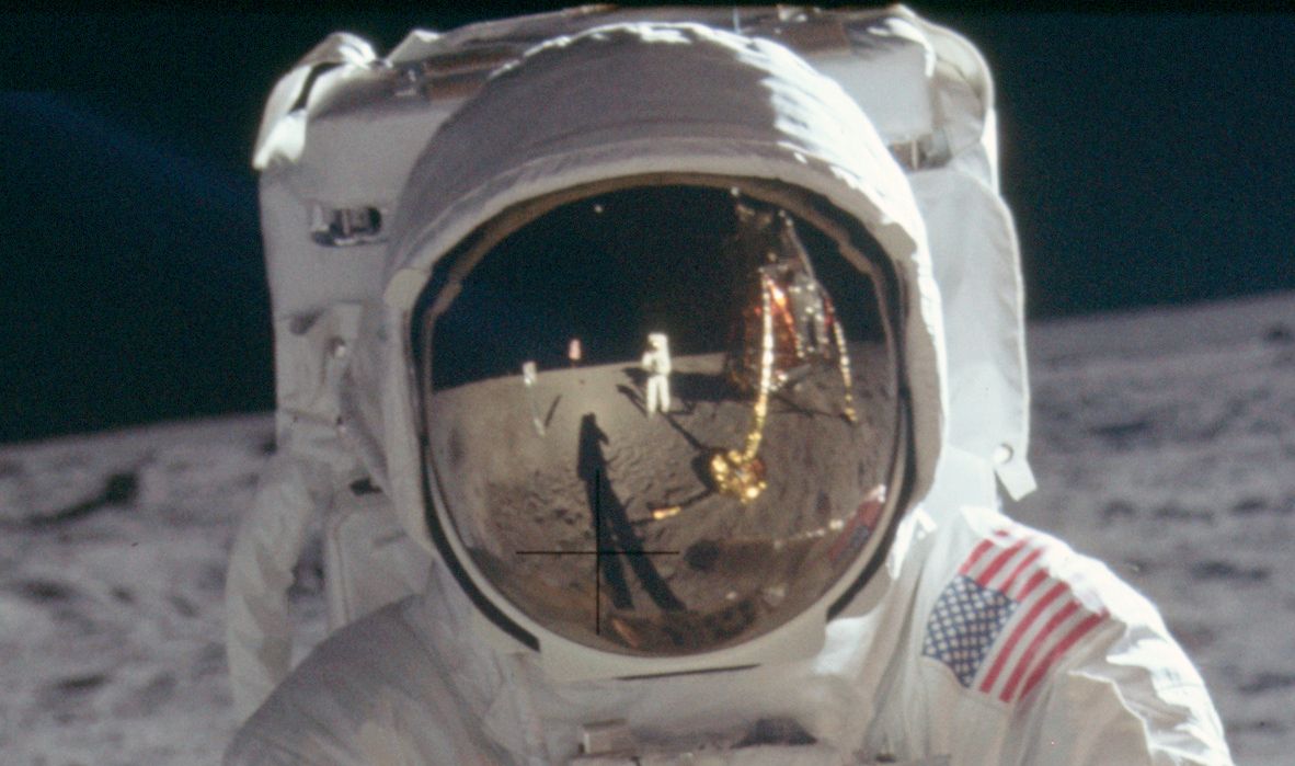 Close-up of an astronaut on the moon, with a reflection of another astronaut and the lunar module in the visor.