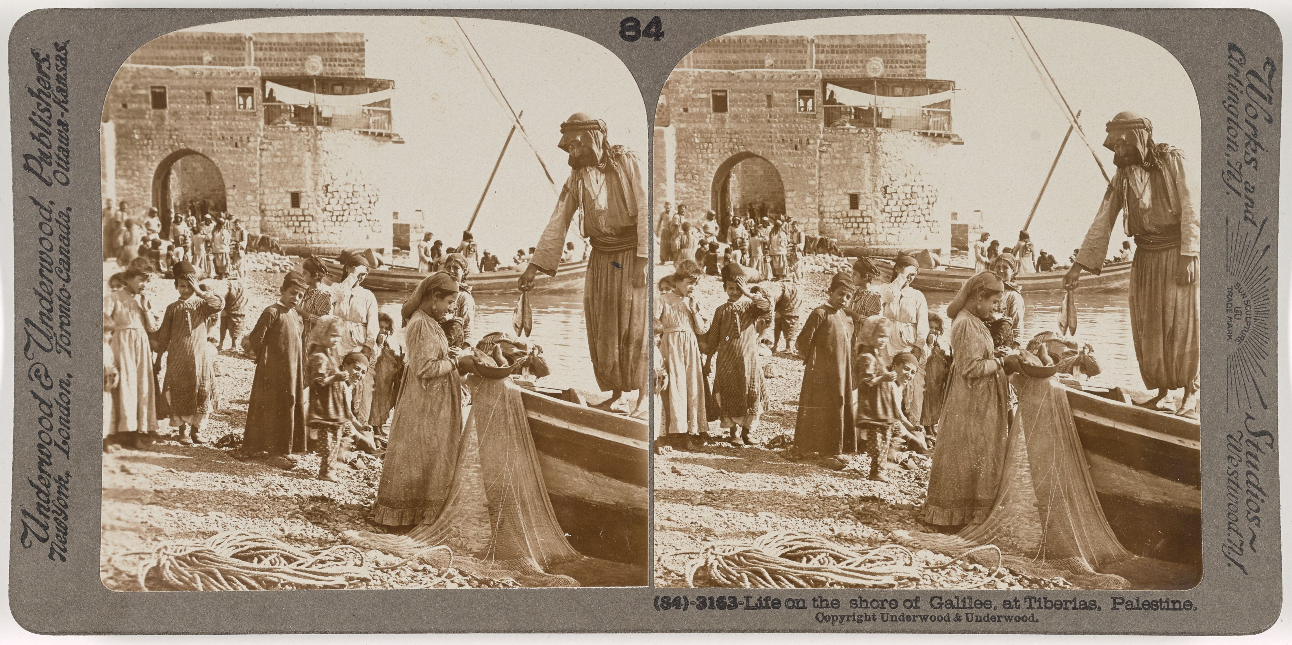 Stereoscopic photograph of life on the shore of Galilee at Tiberias, Palestine. A group of people are near the water's edge, with men in a boat and women on the shore. Buildings and structures line the shoreline in the background.