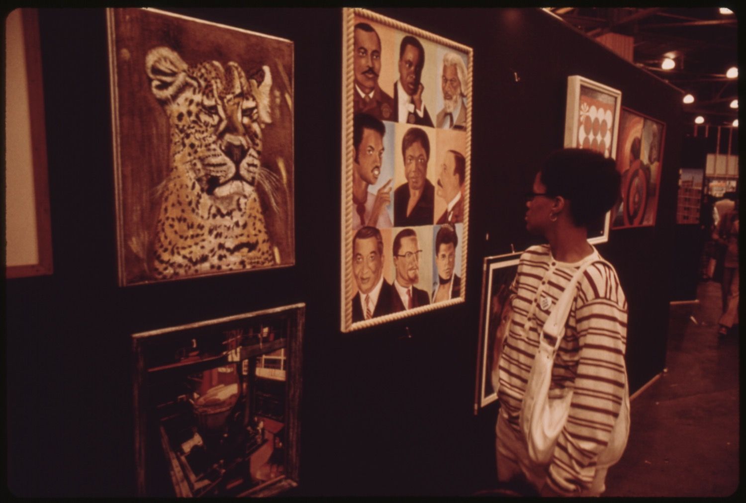 A person in a striped shirt stands in an art gallery, looking at a collection of framed artwork, including a painting of a leopard and a grid of portraits.