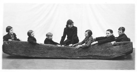 A black and white photograph of seven children and one adult seated inside a long, narrow wooden boat.