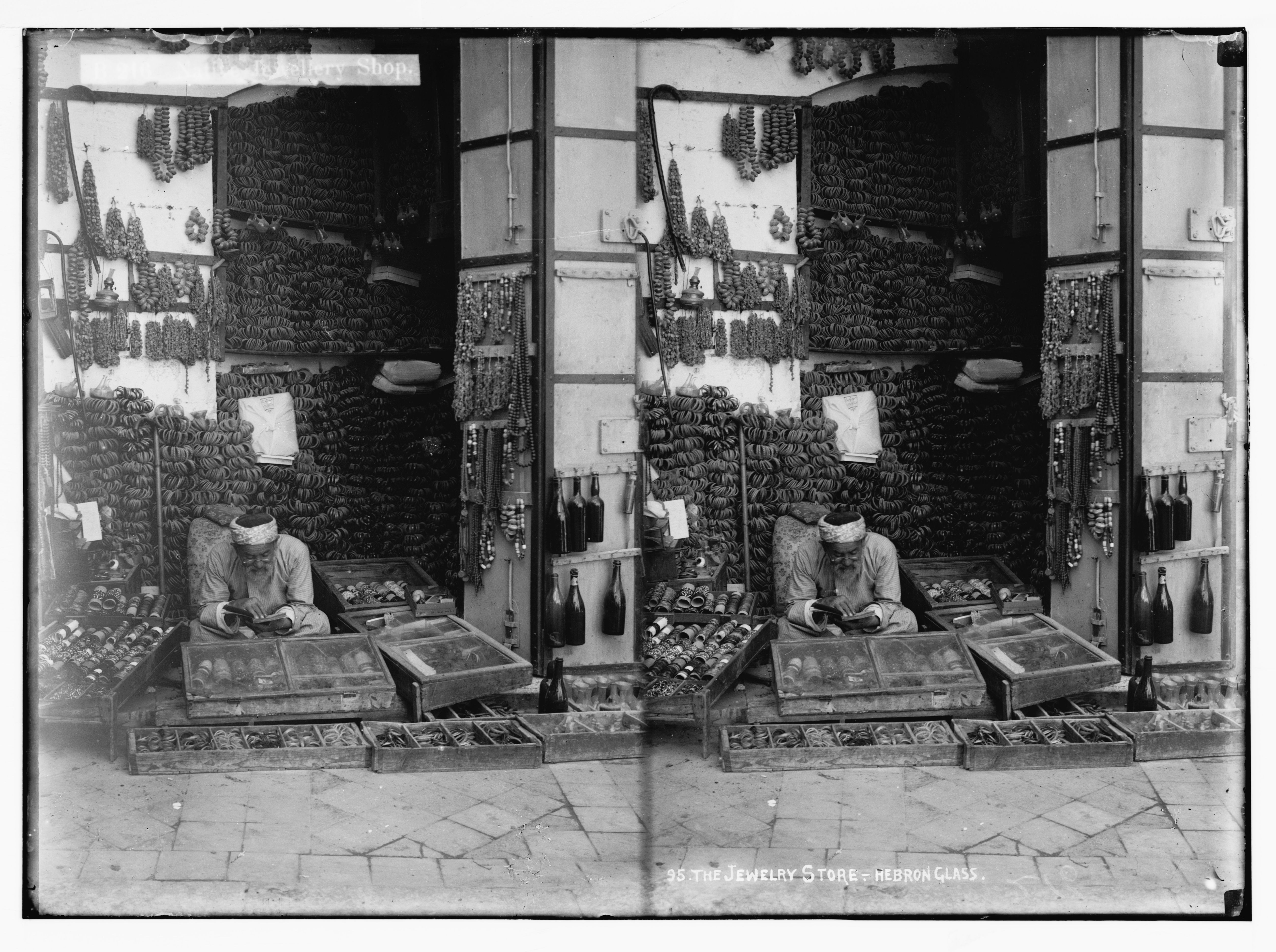Stereoscopic photograph of a jewelry store in Hebron, filled with strings of beads and a man working at a counter.