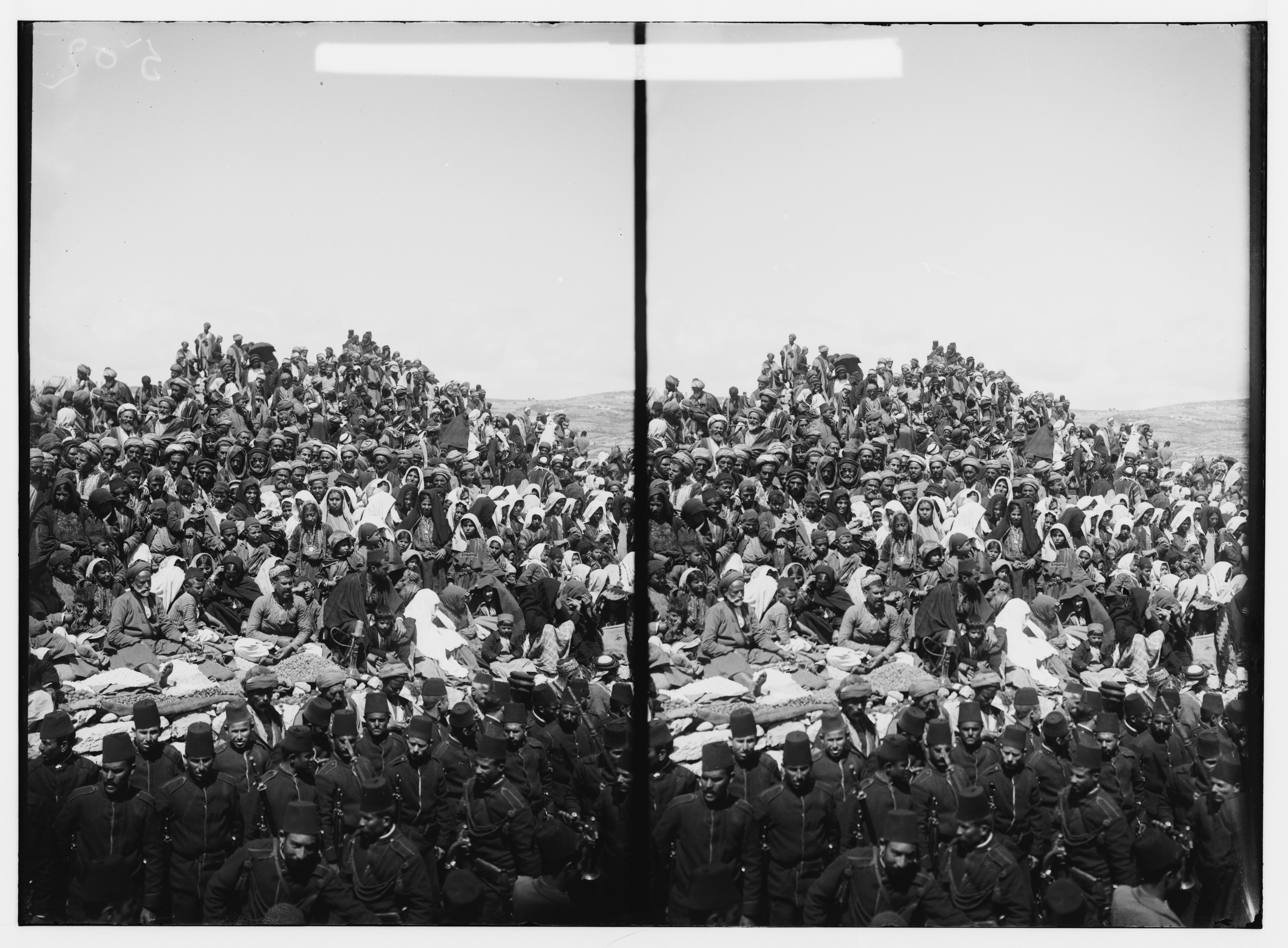 Stereoscopic photograph of a large group of people gathered on a hillside, wearing hats and uniforms, with a barren landscape in the background.