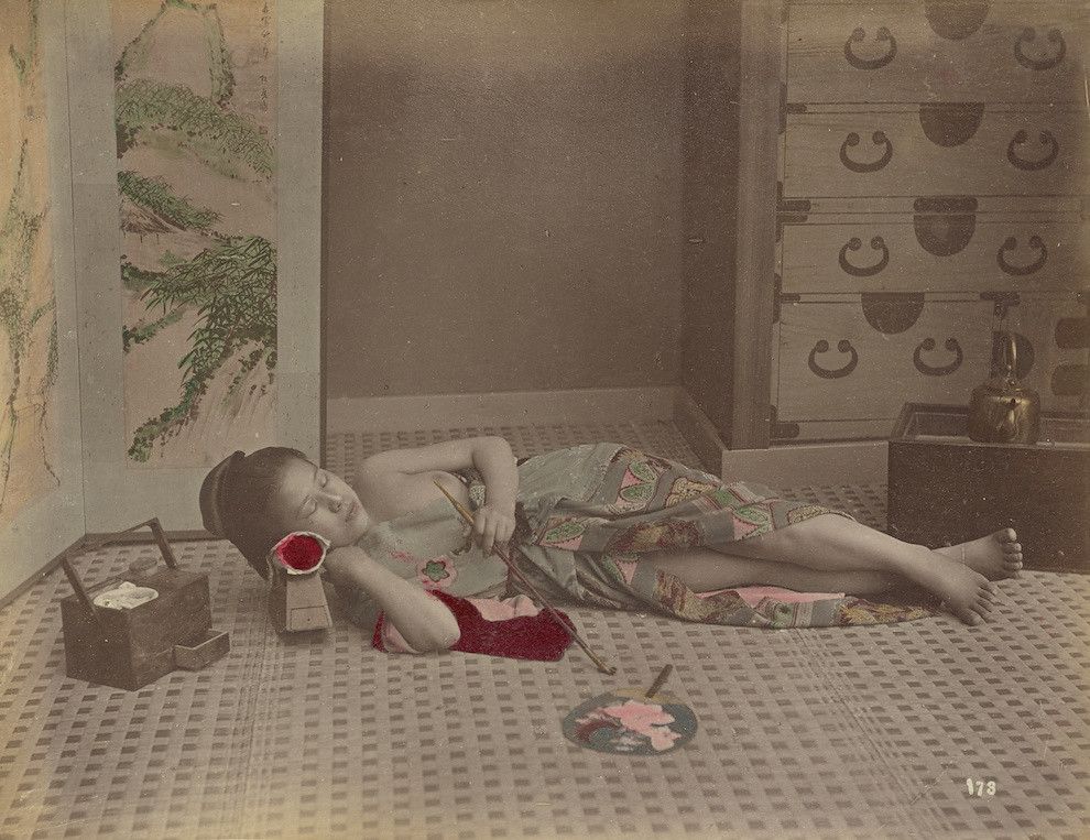 A hand-colored albumen print photograph of a young Japanese woman lying on a tatami mat floor, writing or painting with artistic tools around her.