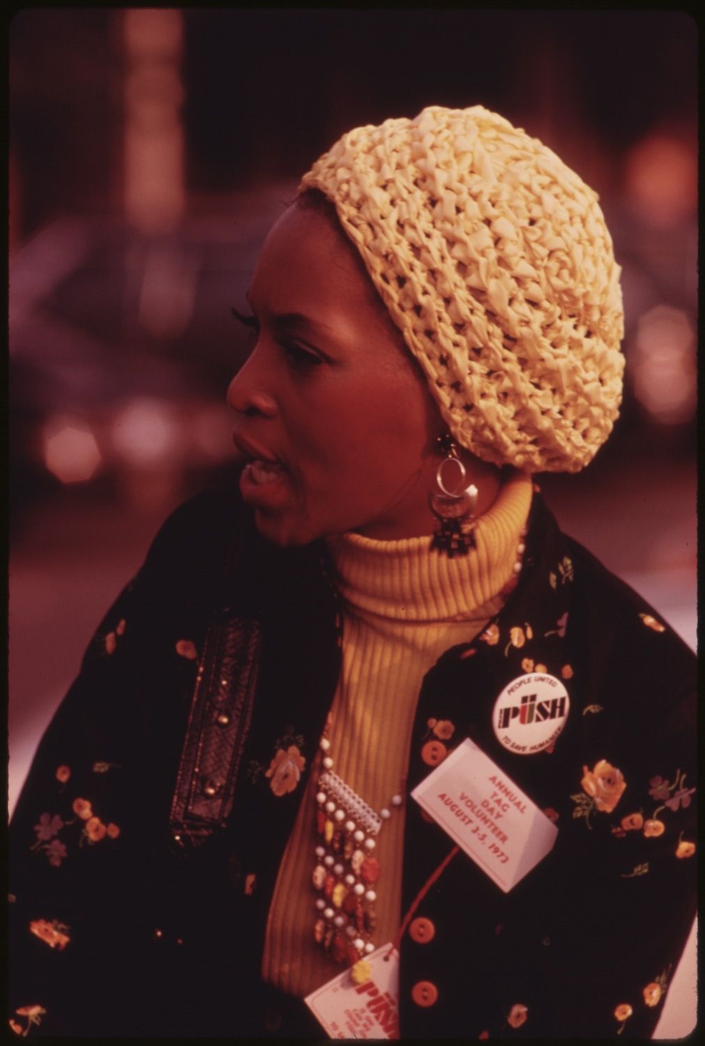 Close-up portrait of a Black woman wearing a crocheted hat, yellow turtleneck, and black floral jacket with 'PUSH' and 'ANNUAL PUSH VOLUNTEER' badges pinned on.