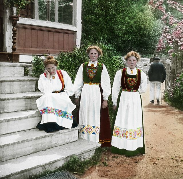 Three women in traditional Norwegian folk costumes pose on stone steps in front of a building surrounded by greenery.