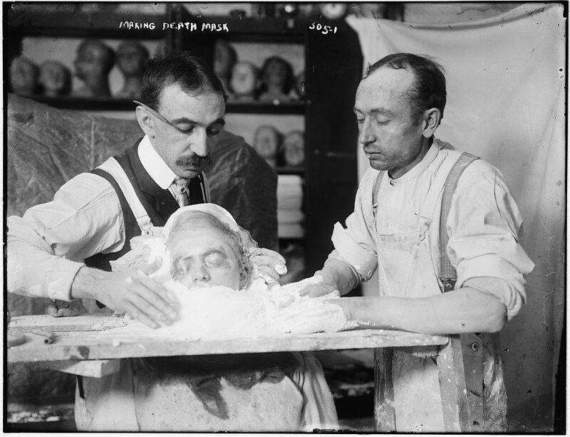 A black and white photograph of two men making a death mask. One man is smoothing plaster around the face of a cast, while the other assists. The scene is somber and clinical, likely taking place in a studio or workshop.