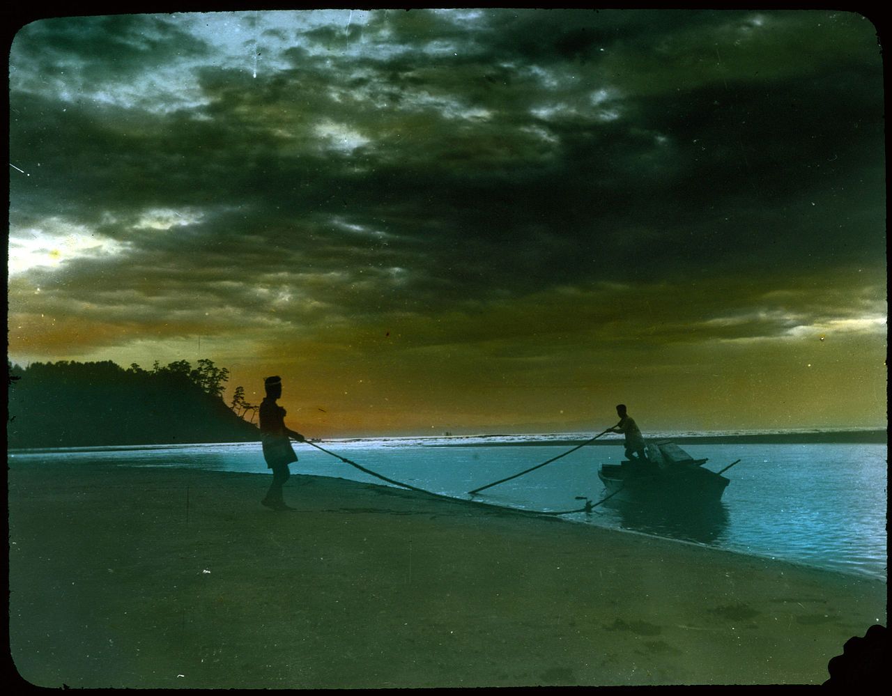 A vintage-style photograph of two silhouetted figures pulling a small boat onto a sandy beach at dusk or dawn. The sky is filled with dark, dramatic clouds, and the water is calm and reflective.