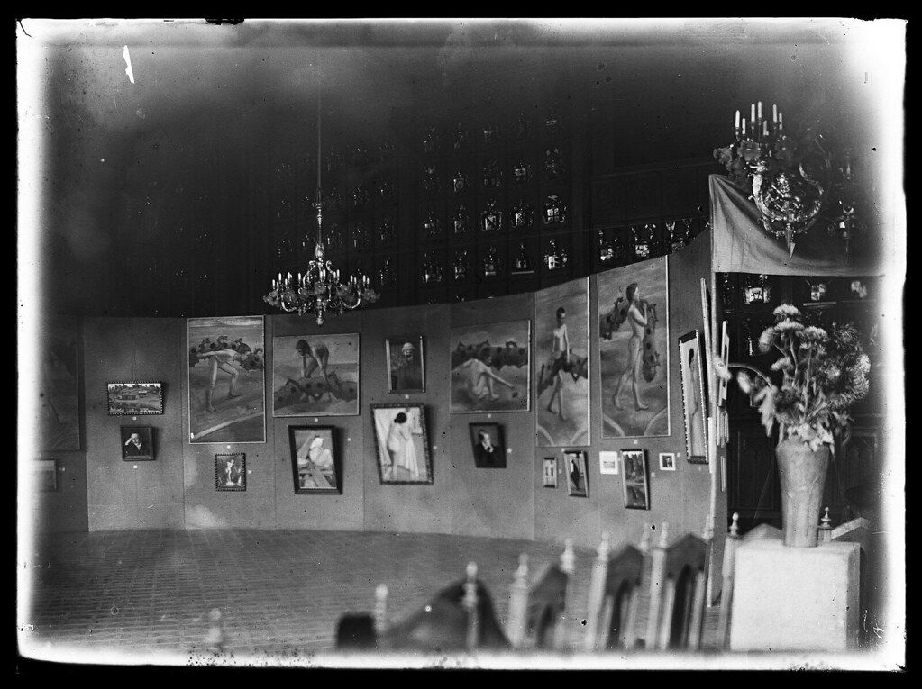 Black and white photograph of an art exhibition with paintings on the walls, a chandelier, and people viewing the art.