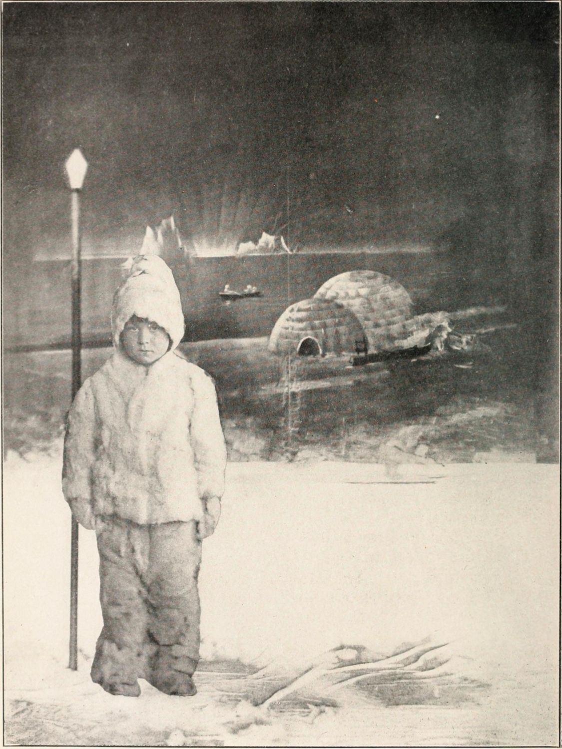 A vintage black and white photograph of a child in winter clothing standing in front of igloos and a snowy landscape.