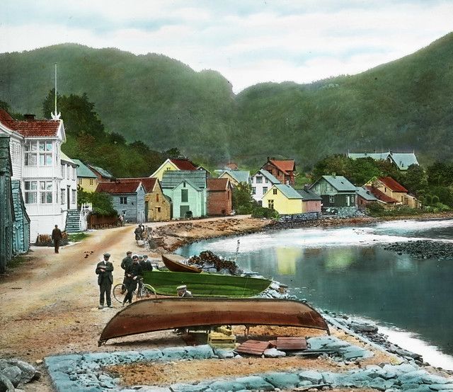 A vintage photograph of a coastal town with colorful wooden houses, boats on the shore, and green hills in the background.