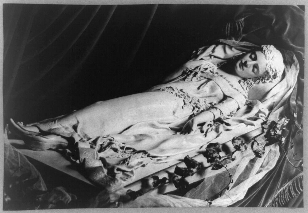 A black and white photograph of a woman lying down in a long, ornate gown covered in flower petals.