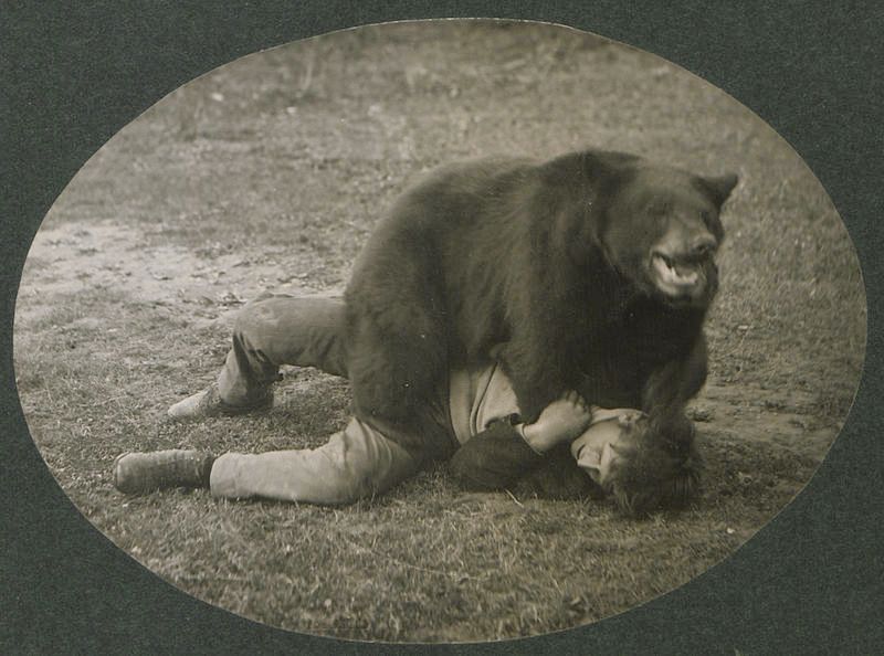 A sepia-toned photograph of a man wrestling with a bear on a grassy field.