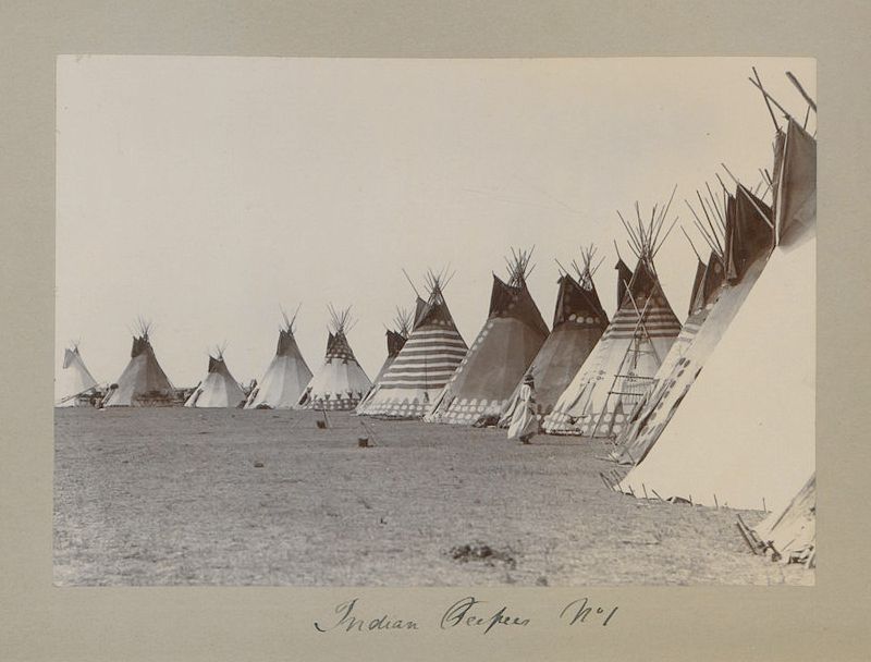 A sepia-toned vintage photograph of a large encampment of teepees in a vast, flat grassland.