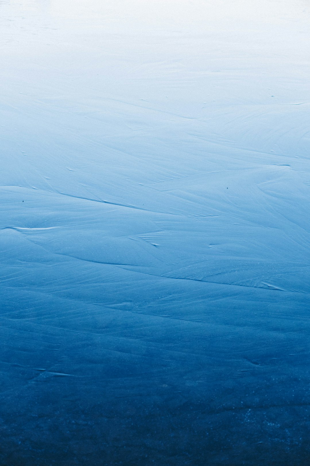 The image shows a surface of ice, likely a frozen body of water. The color palette is dominated by shades of blue, transitioning from a lighter, almost white-blue at the top to a deep, dark blue at the bottom. This creates a gradient effect, suggesting depth or the angle of light hitting the ice.
Subtle textures and patterns are visible on the ice surface. There are faint, meandering lines that resemble cracks or ripples frozen into the ice. These lines are more prominent in the middle of the image and become less visible towards the top and bottom. The overall effect is one of serene coldness and a smooth, expansive surface. The image is minimalistic and focuses on the texture and color of the ice.