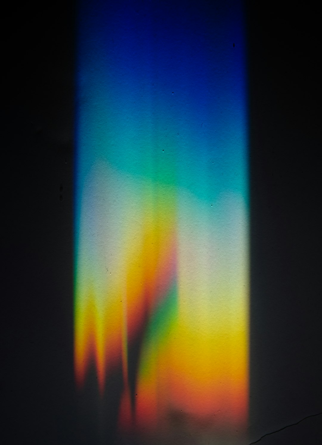 The image features a vibrant and abstract color gradient against a stark black background. The gradient transitions from a deep blue at the top, through shades of green and yellow, and finally to a warm orange and red at the bottom. The colors aren't sharply defined but blend into each other, creating a soft, ethereal effect. There's a sense of light emanating from within the gradient, particularly in the yellow and green areas, giving it a luminous quality. The overall impression is one of fluidity and movement, resembling a stylized aurora borealis or a colorful light leak. The texture appears slightly grainy, adding to the artistic and dreamlike quality of the image.