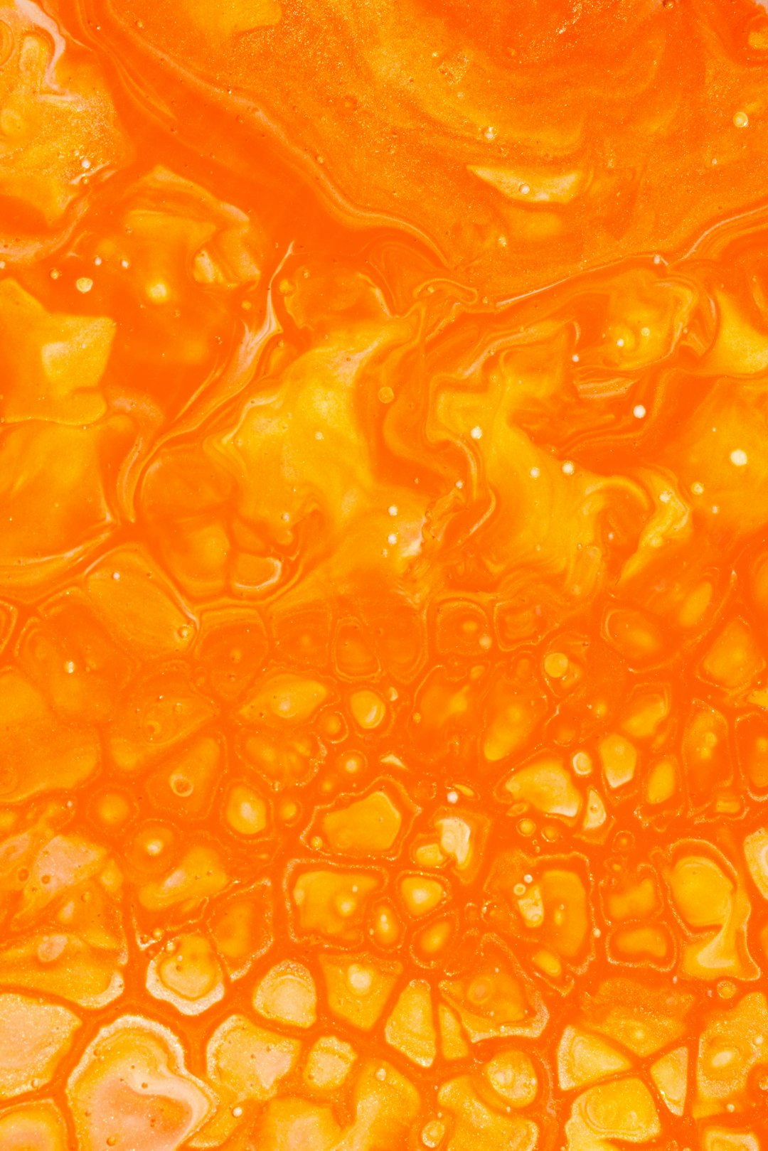 The image is a close-up, abstract view of a vibrant orange fluid. It appears to be a mixture of liquids, possibly paint or dye in water, creating a mesmerizing, marbled effect. The fluid is characterized by a network of swirling patterns and numerous small, irregular bubbles that create a textured surface. The color is a rich, warm orange, with variations in tone that add depth and dimension. The overall impression is one of movement and fluidity, resembling a microscopic view of a colorful, organic substance.