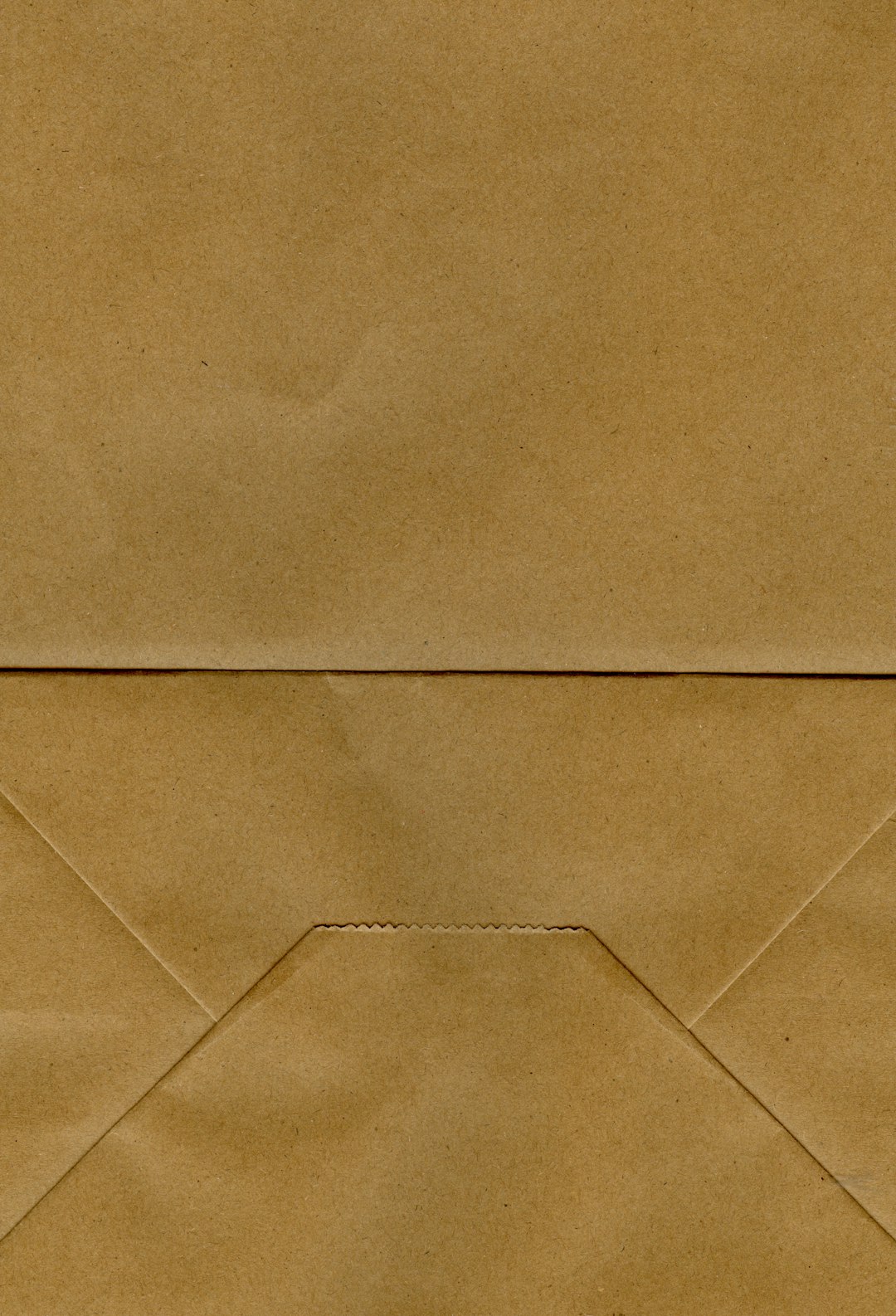 The image shows a close-up of a brown paper bag. The bag is a solid, uniform shade of brown with a slightly textured surface. There's a horizontal crease across the middle of the image, and a vertical crease running down the center. The bottom of the bag forms a triangular shape, with a small, jagged line indicating where the bottom is folded and sealed. The overall impression is one of simplicity and natural texture.