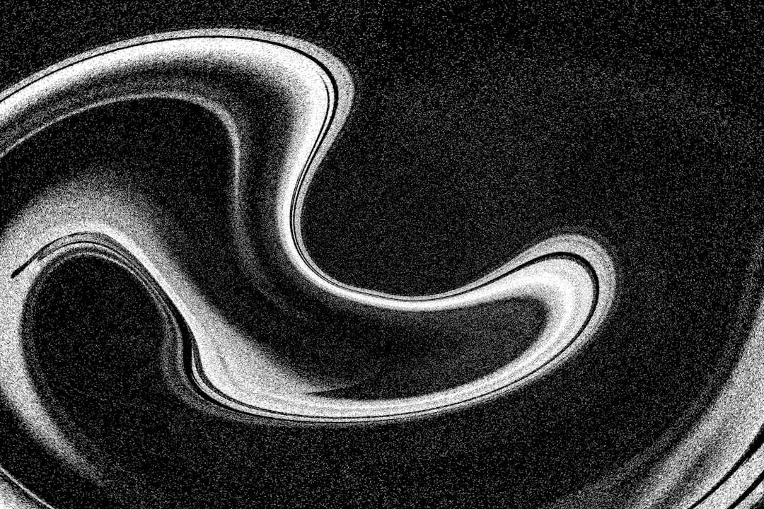 The image is a black and white abstract composition featuring a swirling, ribbon-like shape. The shape is bright white and appears to be made of a continuous, flowing line that loops and curves in on itself. The background is a deep black, and the entire image is covered in a grainy texture, giving it a vintage or distressed look. The contrast between the white shape and the black background is strong, making the swirl stand out prominently. The overall impression is one of movement and fluidity, with a slightly chaotic yet elegant aesthetic.