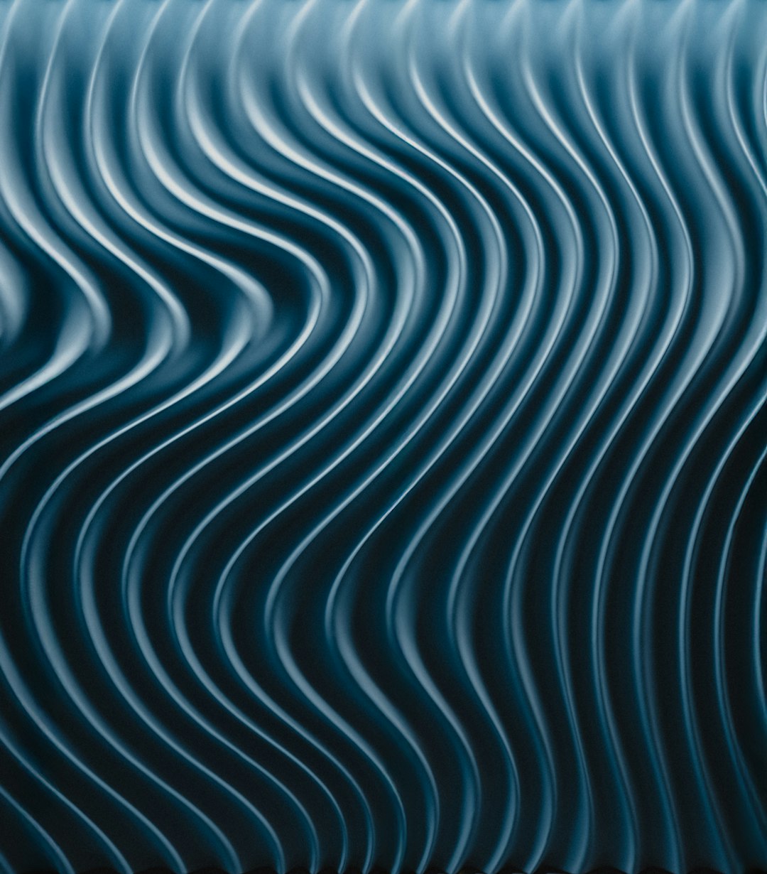 The image presents a close-up of a textured surface with a repeating wavy pattern. The waves are vertical and appear to be formed by a series of closely spaced, undulating lines. The color palette is dominated by shades of teal and dark blue, creating a cool and somewhat mysterious aesthetic. The lighting emphasizes the curves and contours of the waves, with highlights and shadows adding depth and dimension. The overall effect is visually striking and evokes a sense of movement and fluidity.