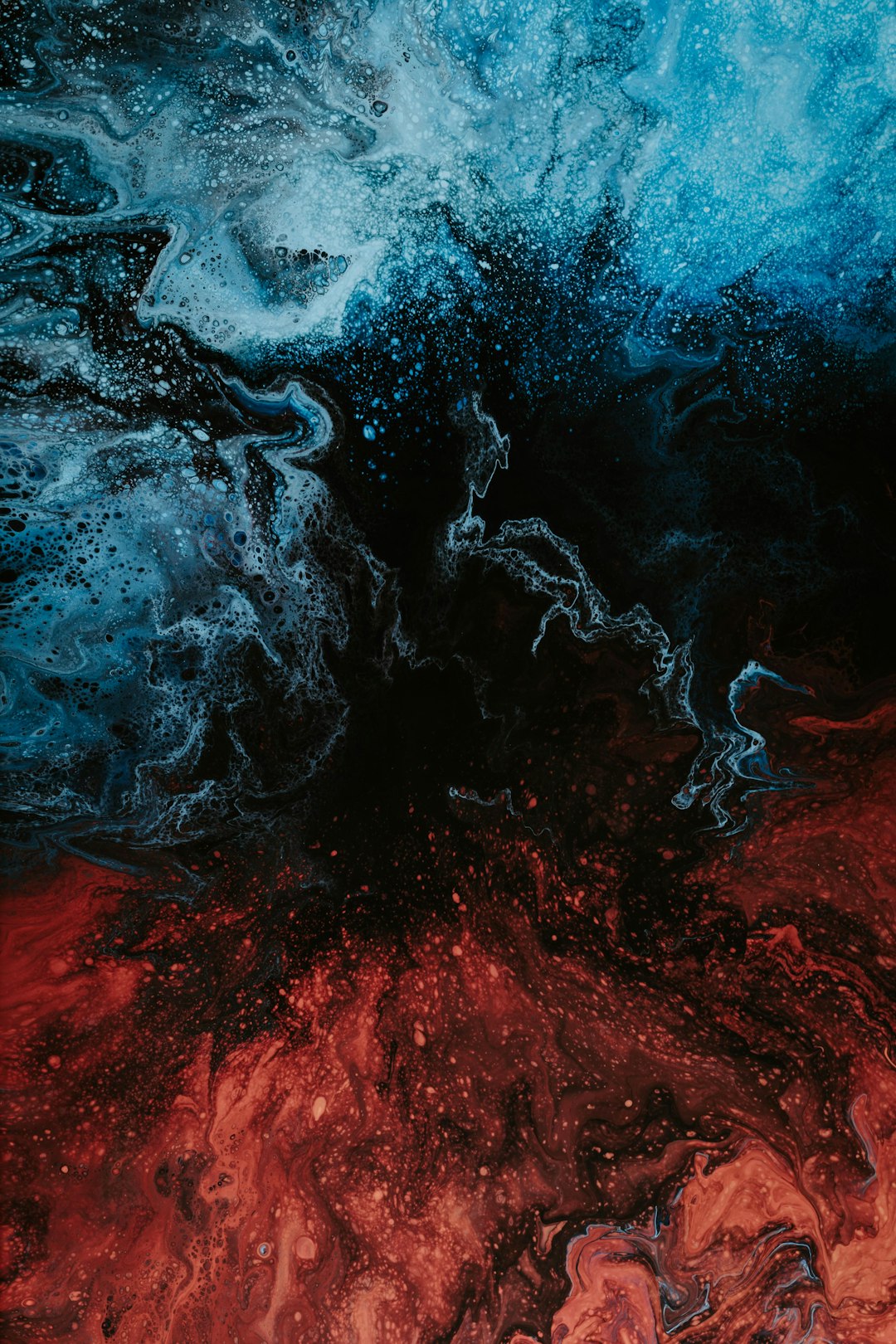 The image is a close-up of an abstract fluid art piece. It features a dramatic contrast between deep blues and fiery reds, creating a visually striking composition. The colors are not cleanly separated but rather swirl and blend into each other, with intricate patterns of veins and tendrils of color weaving throughout. The texture is rich and complex, with numerous small bubbles and irregularities adding depth and dimension. The overall effect is reminiscent of a cosmic nebula or a turbulent underwater scene. The image is dominated by dark tones, with the blues and reds appearing almost luminous against the black background. The composition is dynamic and energetic, with the swirling patterns creating a sense of movement and flow.